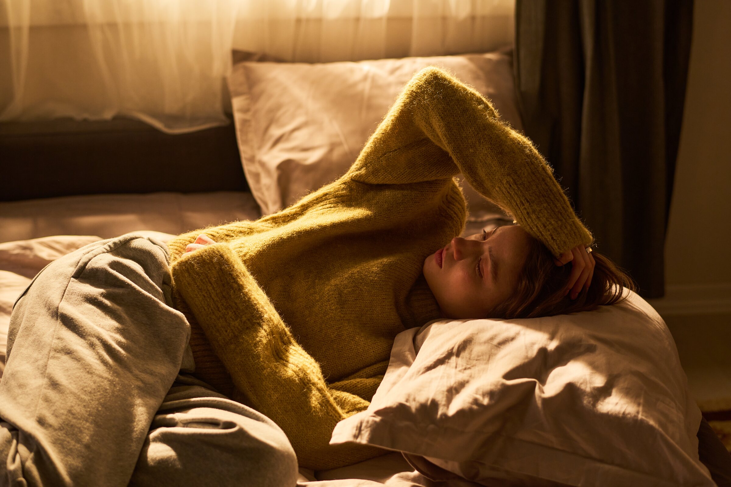 A woman rests in bed in warm afternoon light, one arm over her head
