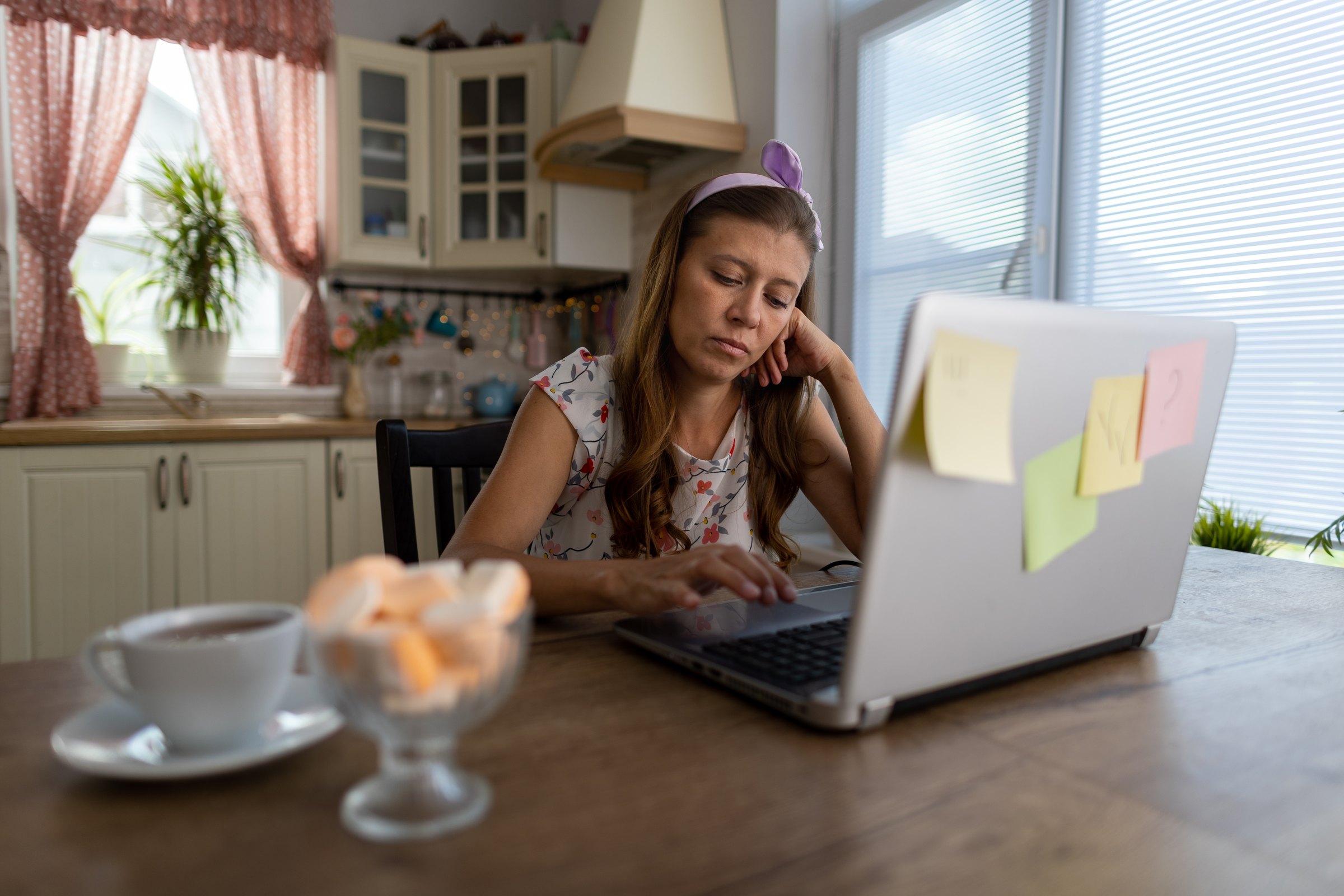 A woman at her kitchen table with sticky notes on her laptop screen, resting her head on her hand.