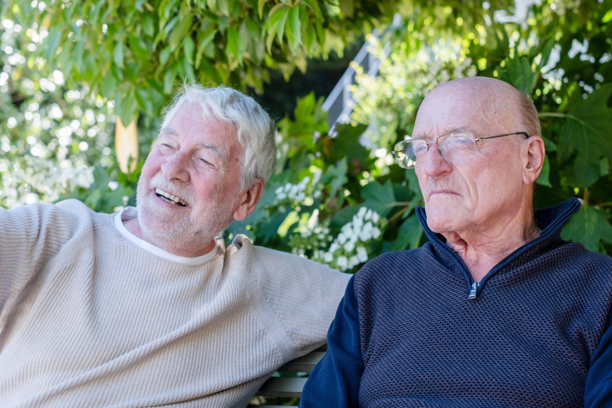Two older men sitting together on a garden bench, one laughing, one listening with his eyes closed.