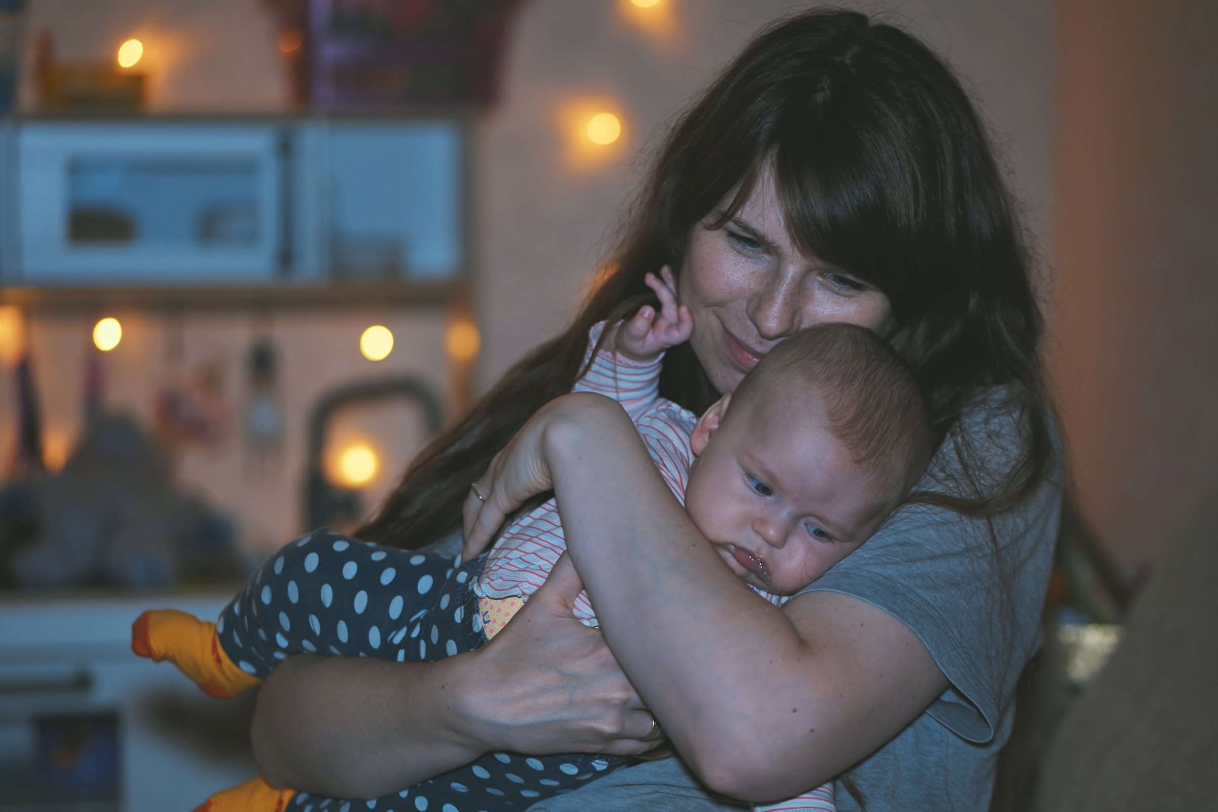 A mother holding her newborn close at night, with soft lights behind them.