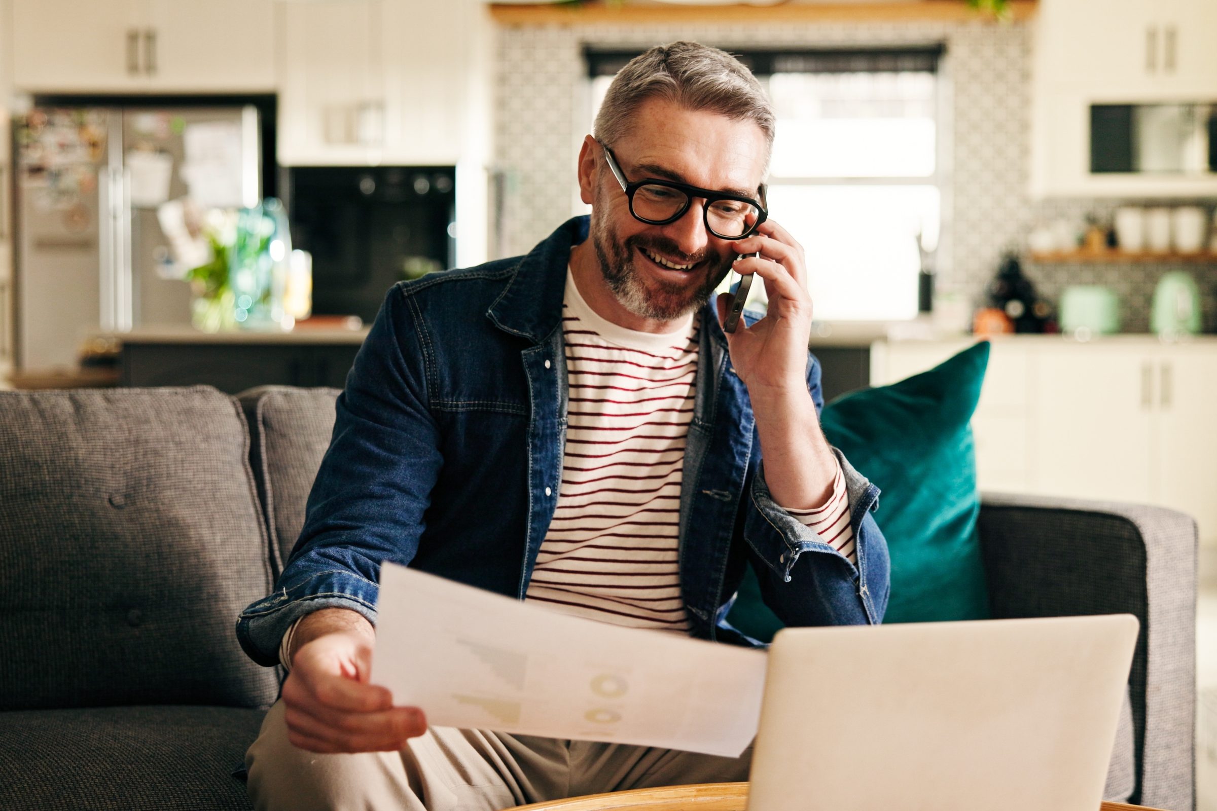 A man on the couch at home smiles on a phone call while reading a document beside his laptop