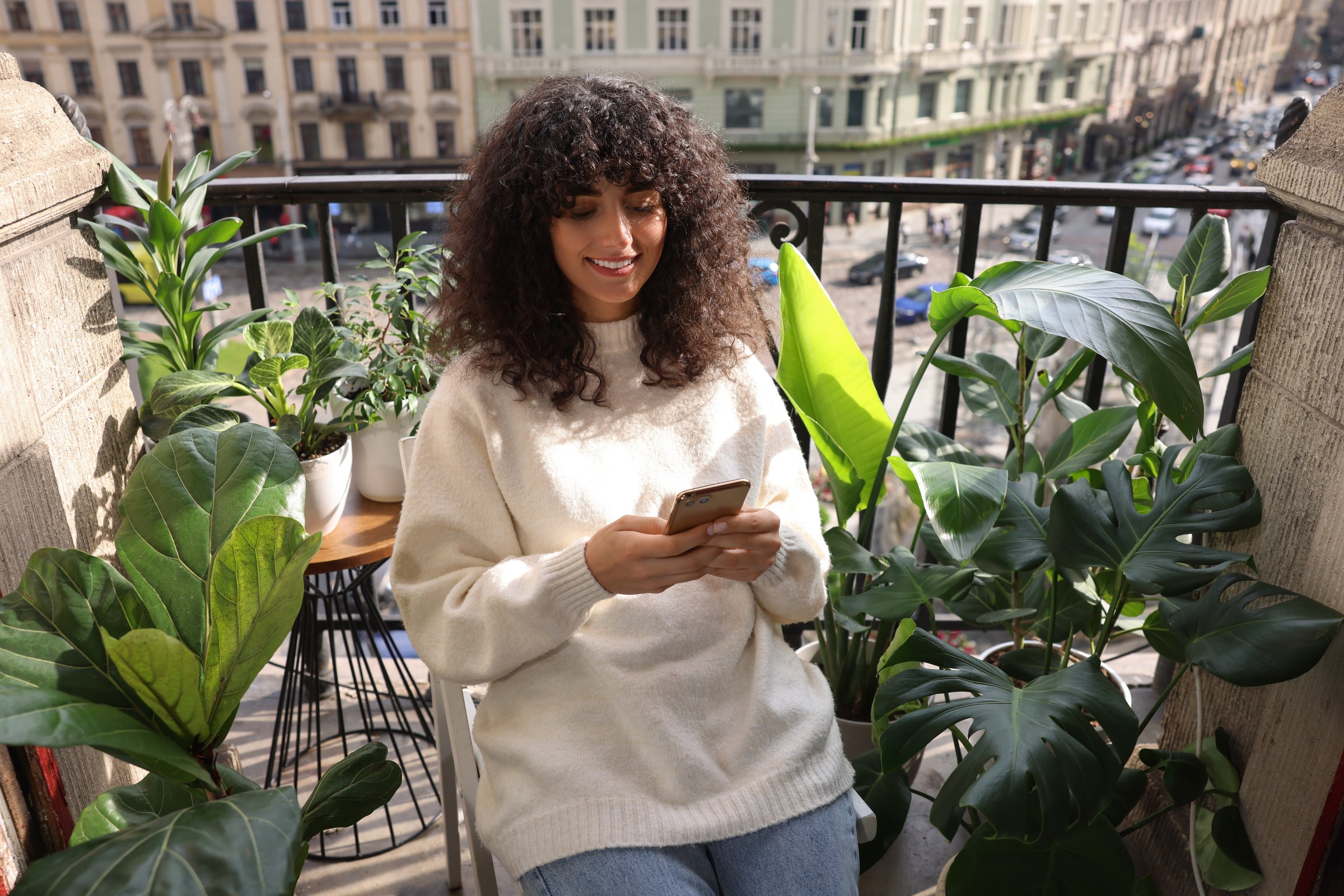A woman sitting among houseplants on a sunny balcony, looking at her phone.