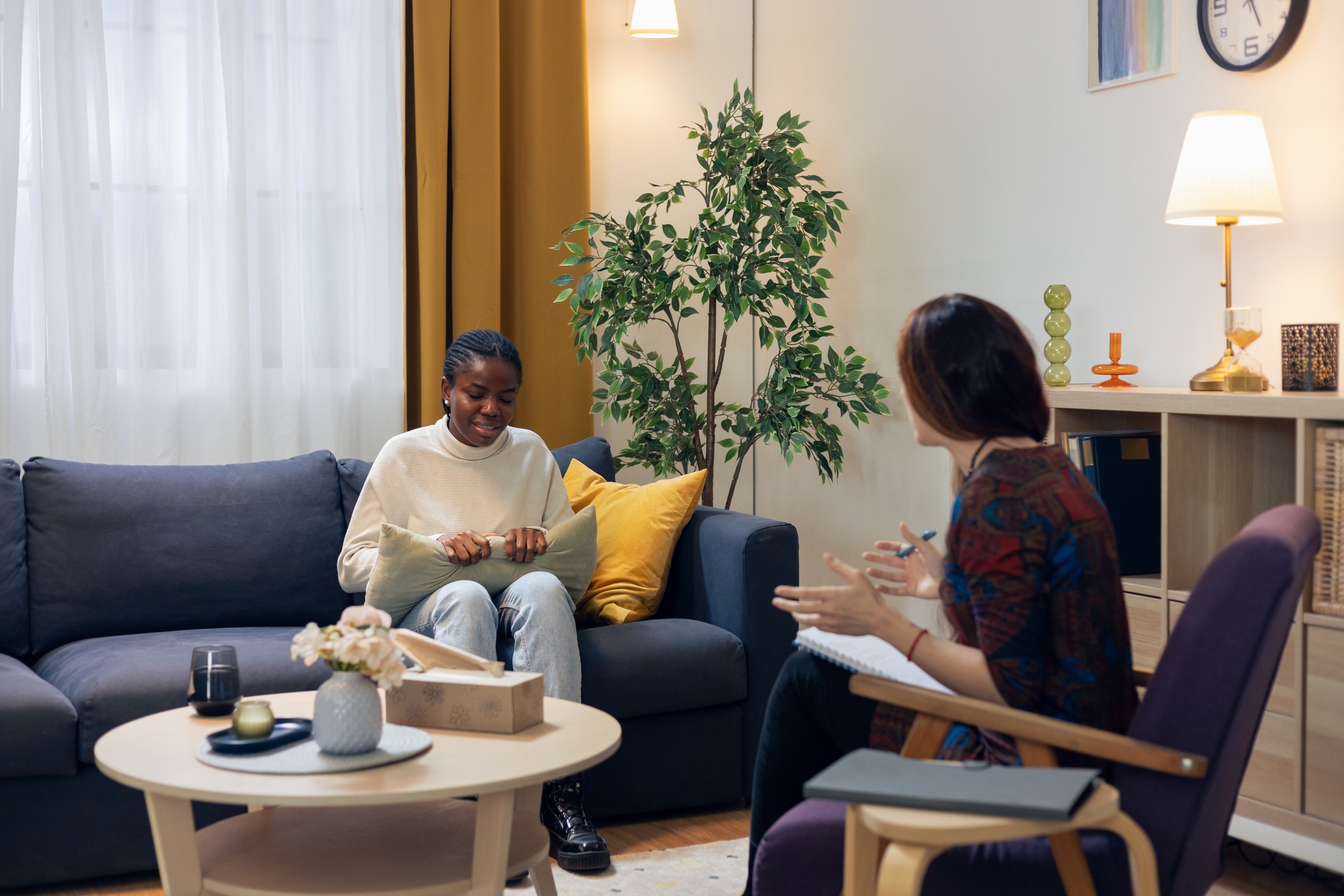 A smiling woman on a couch talking with her therapist in a warm, plant-filled office.